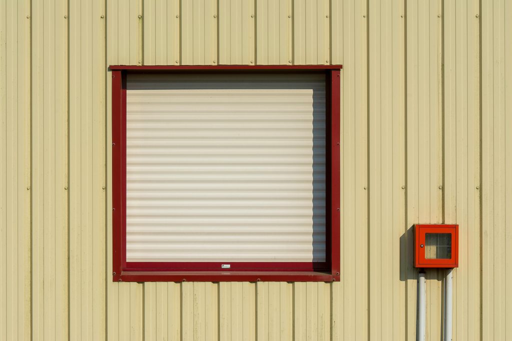 A minimalist industrial building facade featuring a single window with red trim and a small red box.